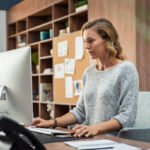 Mature businesswoman working on desktop computer at modern office. Concentrated freelancer finishing project sitting in coworking space using desktop computer. Creative business woman in casual working and sending email.